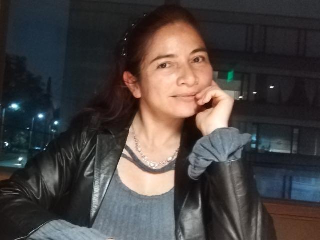 AlessiaRed with dark brown hair, wearing a black leather jacket and gray top, has a silver necklace and a scrunchie on her left wrist, smiling softly.