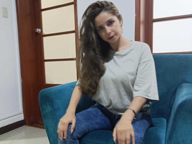SophieTeller with long brown hair, grey t-shirt, and blue jeans, sitting on a teal couch, looking slightly to the side with a subtle smile.