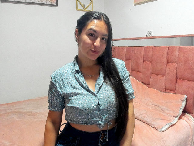 VenusSweetPalmer with long black hair, wearing a blue patterned crop top, showing her small tits, sitting on a pink bed with a pink headboard, smiling softly.
