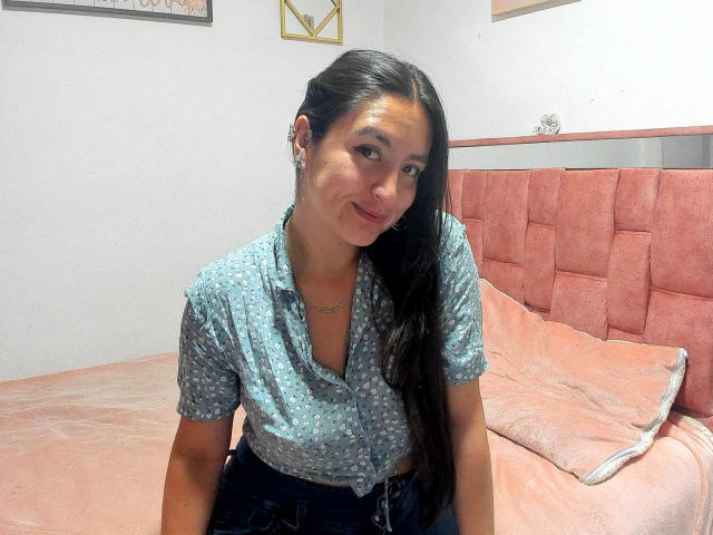 VenusSweetPalmer a de longs cheveux noirs, porte une chemise bleue à motifs, elle sourit, est assise sur un lit rose, et porte un jean foncé, a de petites seins et un collier discret..