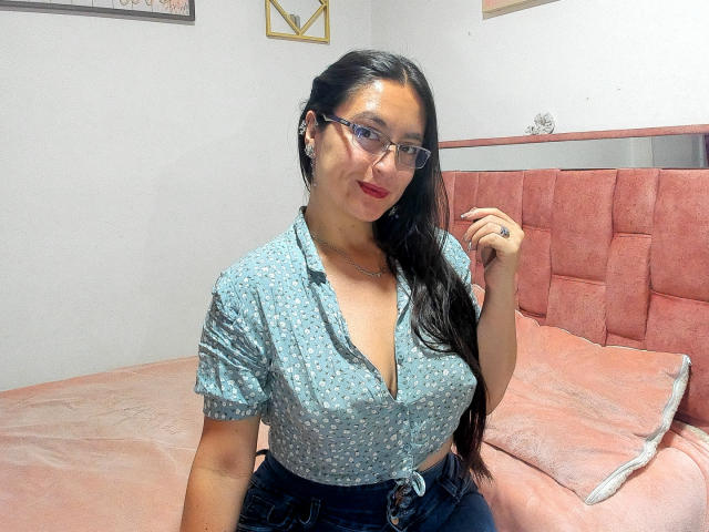 VenusSweetPalmer with long black hair, glasses, and red lipstick, wearing a blue patterned shirt, showing her tits, sitting on a pink bed, hand near face.