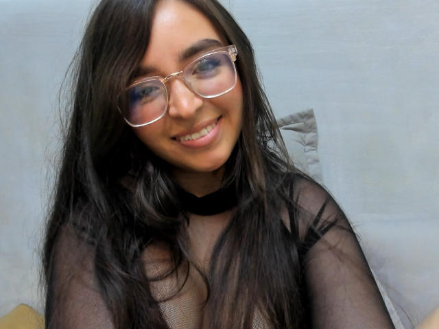 EvaMcAllister with long black hair, wearing clear glasses and a sheer black top, smiling at the camera, showing her tits through the fabric.