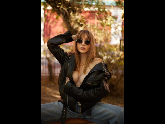 Shezel-hot with round sunglasses, black leather jacket, and exposed boobs, sitting outdoors, hand in hair, wearing blue jeans, gold necklace, and a confident, sexy look.