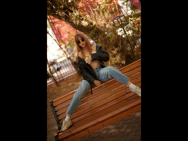 Shezel-hot sits on a wooden bench with legs spread, wearing a black jacket open to expose her tits, blue jeans, and white sneakers, in a park.