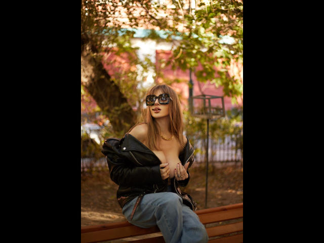 Shezel, con los senos expuestos, vistiendo una chaqueta negra y gafas, sentada en un banco, con jeans, un collar dorado, hojas de otoño, cabello castaño, mirando a la cámara, al aire libre..