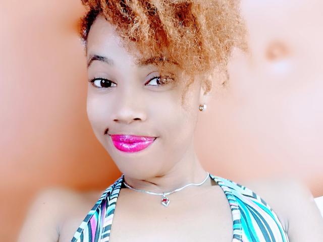 HotEllySexy with curly orange hair, pink glossy lips, brown eyes, small earrings, striped halter top, silver necklace, smiling at the camera, showing her cute face and smooth skin.