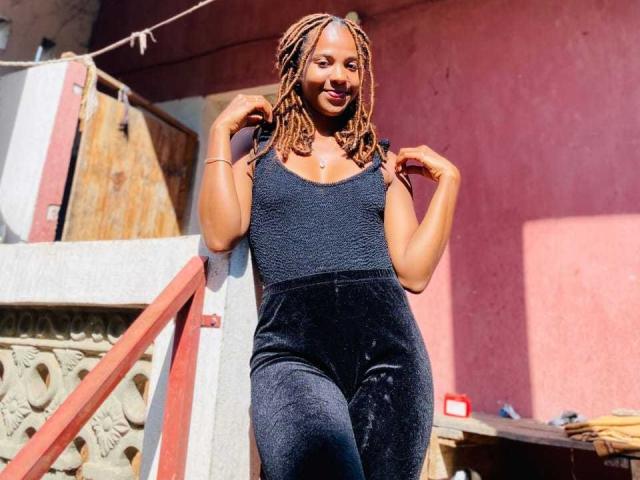 MilSexy with brown braided hair, smiling, wearing a black textured sleeveless top and high-waisted velvet pants, standing near a red and gray railing.