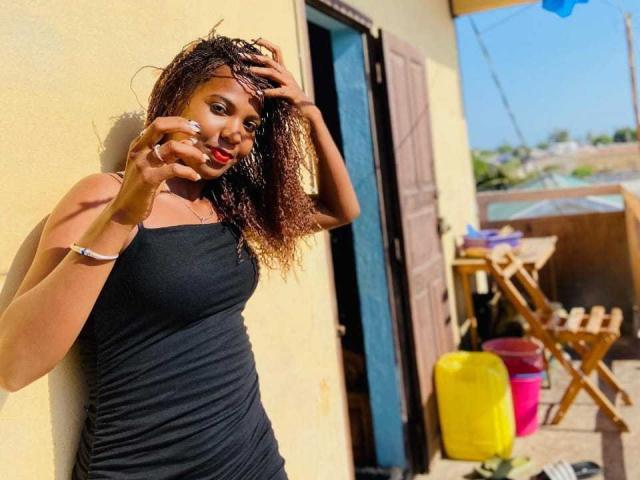 MilSexy, dark-skinned, with curly brown hair, wearing a black dress, touches her head with one hand and her face with the other, standing against a yellow wall.