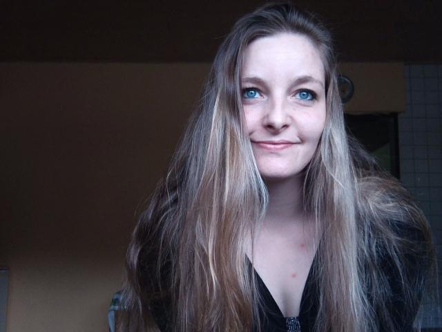Jennylove-hot with long, dirty blonde hair, blue eyes, and a small smile, wearing a black zip-up, showing a bit of cleavage, in a dimly lit room.