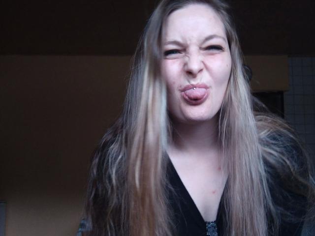 Jennylove-hot with long blonde hair, tongue out, and a small lip piercing, looking goofy with slightly squinted eyes. She's wearing a black top with a furry texture.