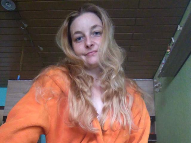Jennylove-hot with long blonde wavy hair, orange fluffy robe, and subtle smile, showing small tits under robe, in a wooden ceiling room.