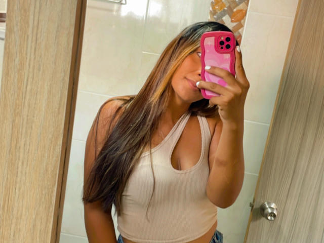 EmelyOwen with long brown hair, pink phone case, and white tank top, showing her tits in a mirror selfie. Her hand covers her mouth, looking cute.
