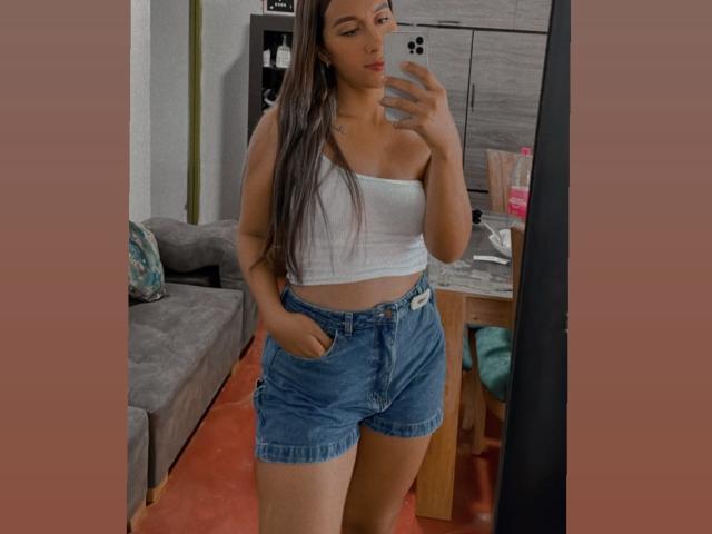 EmelyOwen with long brown hair, white strapless top, and blue denim shorts, standing in front of a mirror, hand in pocket, holding phone, visible butt in mirror reflection.