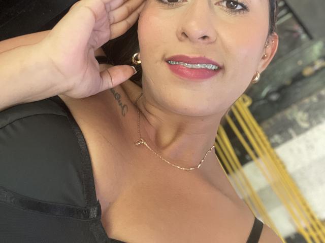 EmelyOwen's face with black bra, silver teeth, gold hoop earrings, black hair, tan skin, necklace, tattoo near collarbone, hand touching cheek, slight smile. Boobs visible in bra.