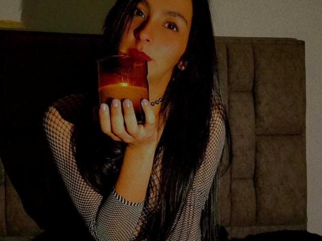 KaatiaRoss with long black hair, fishnet top, purple nail polish, and choker, sipping a dark drink, puckering lips, on a brown sofa, looking at the camera.