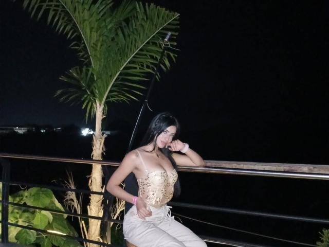 ShantalTurner with black hair, wearing a beige, bejeweled top and beige pants, leaning on a railing, her hand near her mouth, by a tall palm tree at night.