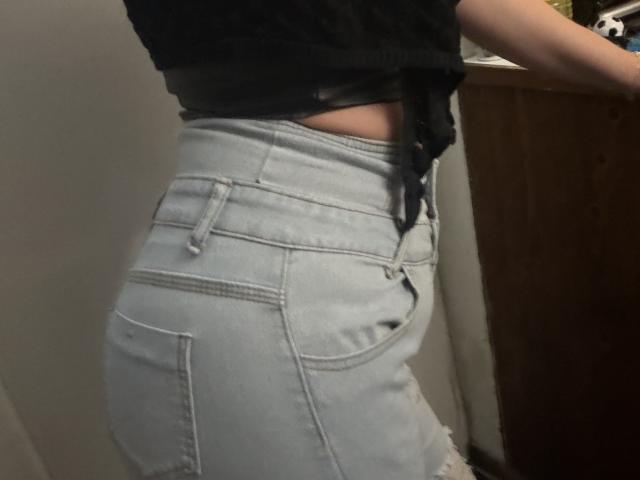 SerenaFr's ass in tight, light blue, ripped denim jeans, showing her buttcheek, with a black top tied above her waist, and her hand on a wooden surface.