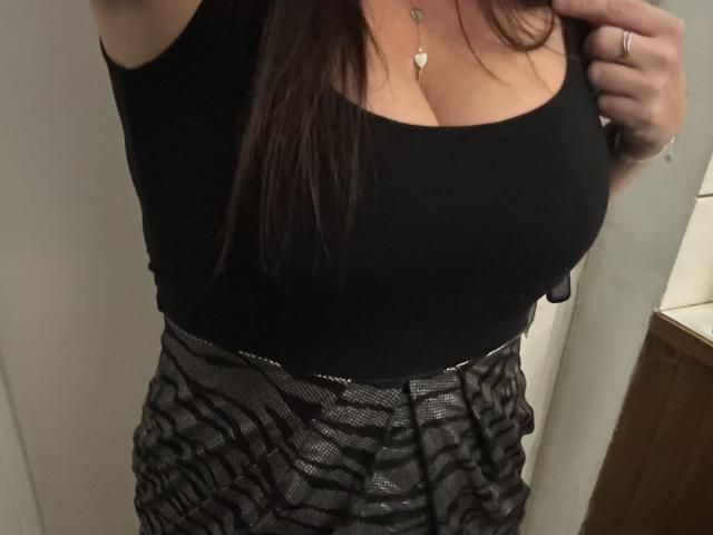 Le débardeur noir de SerenaFr met en valeur sa poitrine généreuse, son décolleté et un collier avec un pendentif en forme de cœur. Elle porte une jupe brillante avec un motif ondulé, et a une main posée sur sa poitrine..