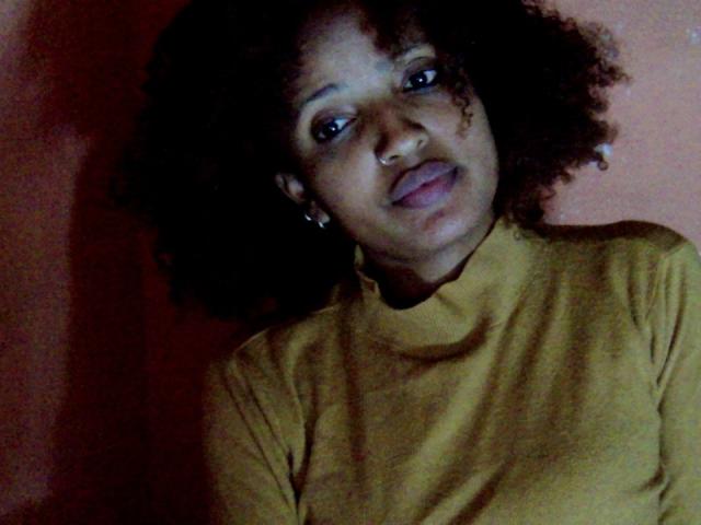 Annaellah, avec ses cheveux noirs et bouclés, porte un pull jaune col roulé et a légèrement sa langue dehors, tout en regardant vers le haut avec un sourire discret. Son nez est percé. L'éclairage est tamisé..