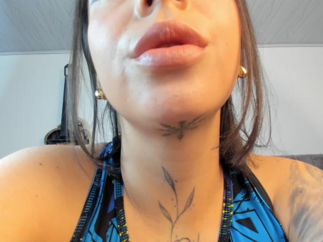 KayleeM's face, showing full lips with a slight pout, a tattoo under her chin, and another tattoo on her neck, wearing a blue and black patterned halter top.