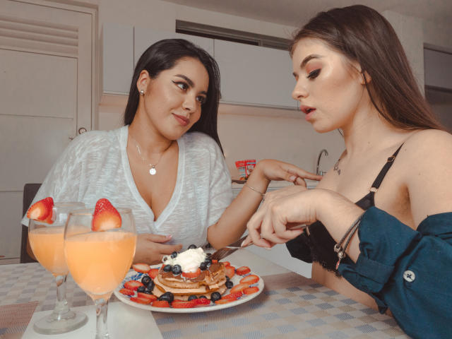 PaolaYKatty with dark hair in a white top, looking at PaolaYKatty with brown hair in a black bra, slicing a fruit-topped pancake, orange drinks with strawberries.