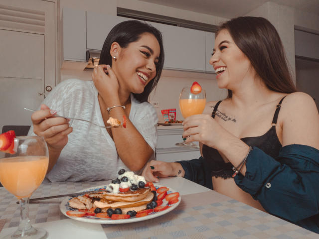 PaolaYKatty with medium tits, black bra, and blue shirt, and another PaolaYKatty with small tits, white shirt, eating pancakes with strawberries, blueberries, and cream, drinking orange cocktails.