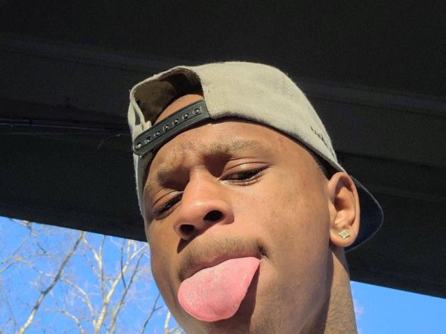 StudTrisO has dark brown skin, a gray baseball cap, and is sticking out his pink tongue with a goofy expression. His ears are pierced with small studs.