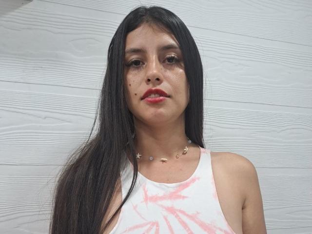 MadissonFontaine with long black hair, wearing a pink and white tie-dye tank top, has small moles on her cheeks, a necklace with small stars, and red lipstick.