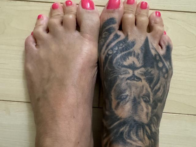 AndraD's feet with pink nail polish, one foot showing a detailed black lion tattoo on the sole, both feet positioned side-by-side on a wooden floor.