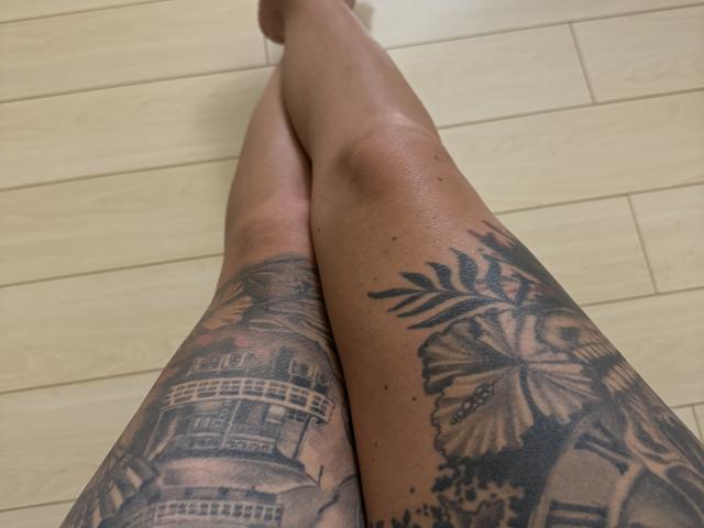 Andras Beine mit detaillierten Tattoos: Das linke Bein zeigt eine TARDIS aus "Doctor Who", das rechte Bein eine große Schmetterling mit Farnen. Holzboden, barfuß..