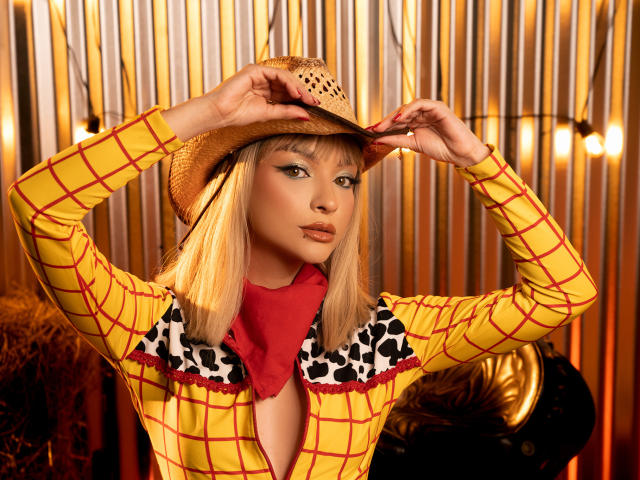 MaryMilan wearing a yellow plaid shirt with black and white cowprint collar, red scarf, and straw hat, holding it on her head, blonde hair, and orange lipstick.