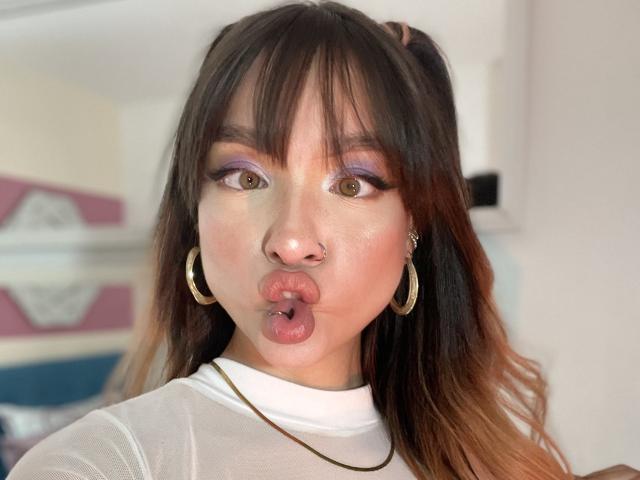 MaryMilan with pouty lips, purple eyeshadow, brown hair in pigtails, gold hoop earrings, white sheer top, and a nose ring, looking directly at the camera.