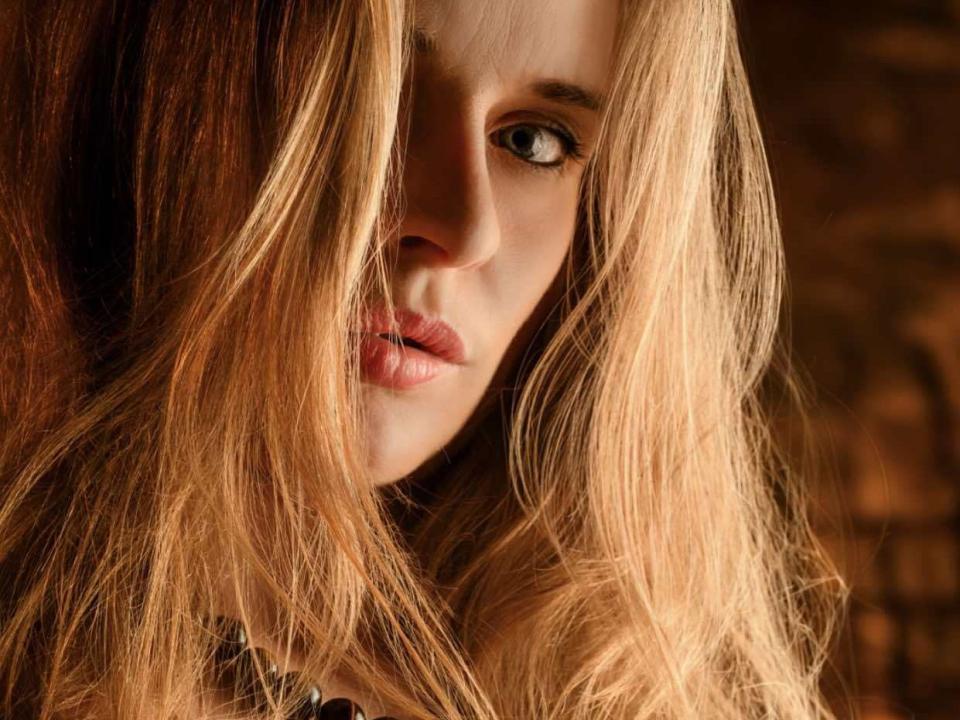XPixieX has long, wavy blonde hair covering one eye, with pinkish lips and a slight pout, in a warm, brown-toned, close-up shot. Her eye is partially visible.