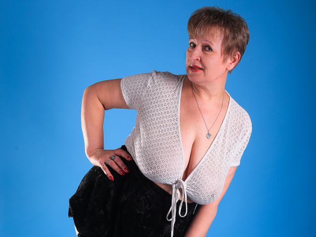 A photo of SugarBoobs with short brown hair, white crochet top showing big tits, black skirt, hand on hip, blue background, looking to the side with a slight smile.