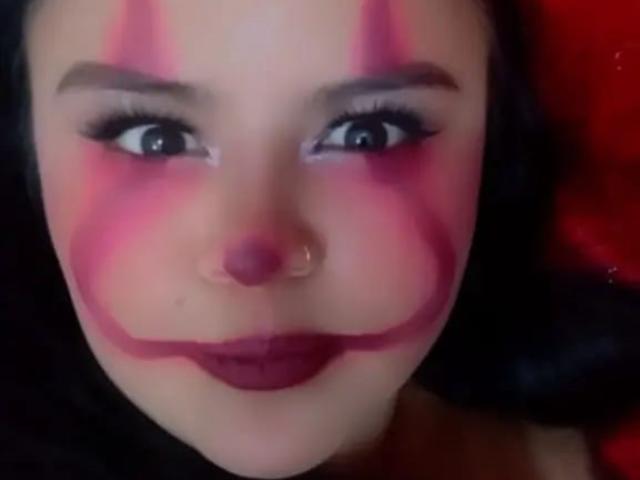 AllizeWalkers has pink clown makeup on her face, with pink stripes on her cheeks and nose, and dark red lipstick. She has a nose ring and dark hair.