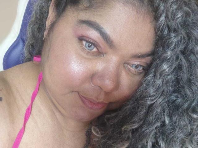 BrunetteBlue with curly gray-black hair, pink eyelid shadow, and pink halter strap, has a subtle smile and slightly raised eyebrows, showing small nose tattoo on left side.