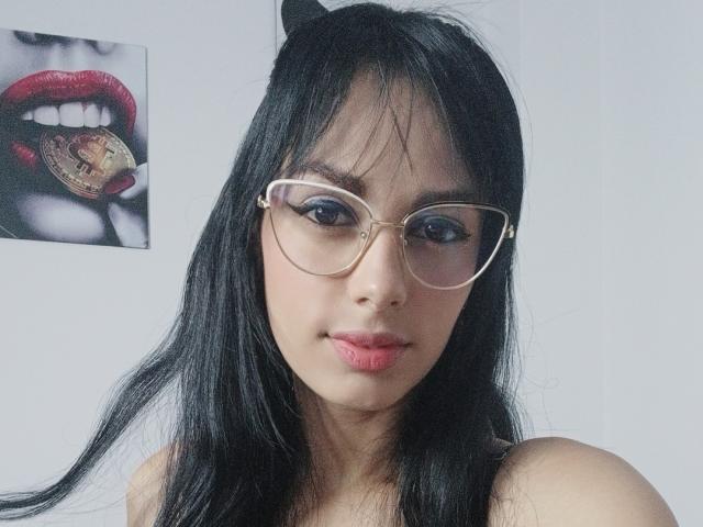 AnaisHilton has black hair, cat ears, and wears clear glasses with dark eyeliner. She has pink lips and a gold ring in her mouth. Her tits are partially visible.
