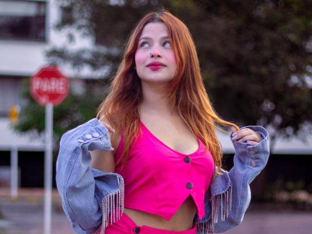 ShantalSummer with red hair, pink top, and buttoned chest, wearing an unbuttoned denim jacket, looking up with pink lipstick and a slight smile, holding jacket sleeves.