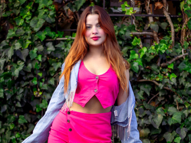 Shantal, met lang rood haar, een roze topje en een bijpassende roze rok, draagt een blauwe jas met franjes en staat voor een groene, bladerrijke achtergrond. Haar borsten zijn zichtbaar onder de top..