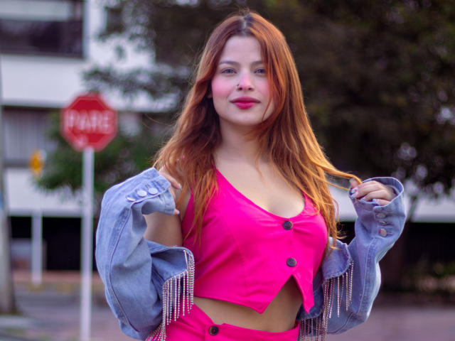 ShantalSummer con una blusa rosa y roja, mostrando los pezones en un sujetador rosa, una chaqueta vaquera colgando de los hombros, cabello castaño largo, lápiz labial rosa, de pie frente a una señal de stop borrosa..