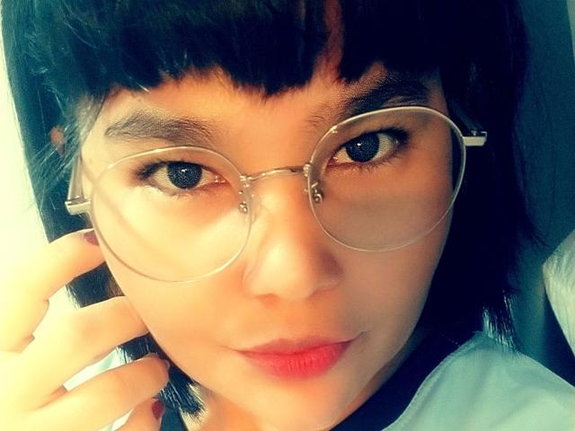 PrittyAlicia with round glasses, dark straight hair, and red lipstick, staring into the camera with blue eyes. Her hand touches her ear. She's cute and slightly smiling.