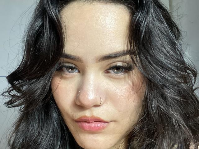 SammyColleman a les cheveux noirs et ondulés, un piercing au nez et des lèvres roses avec un léger sourire. Elle regarde l'appareil photo avec ses yeux verts et un maquillage discret..