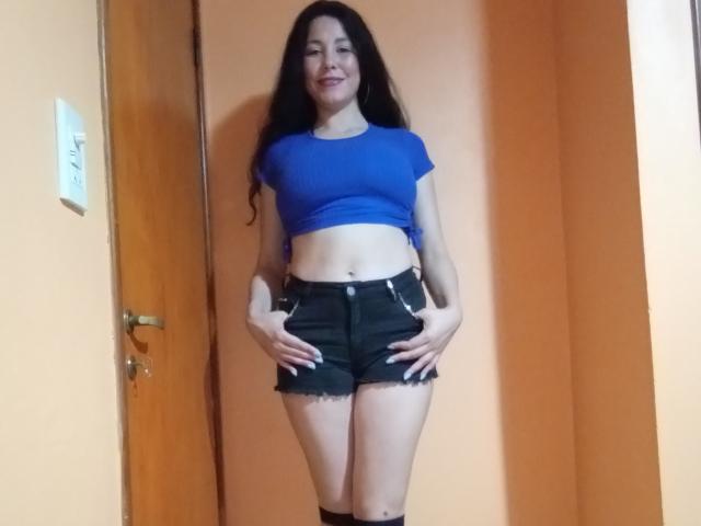 Satika with long black hair, blue crop top, black frayed shorts, hands on her thighs, smiling, standing in a room with orange walls and a wooden door.