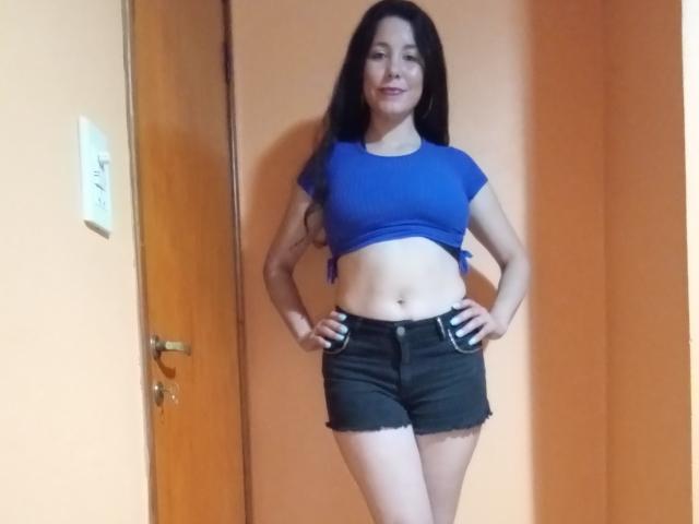 Satika has long black hair, a blue crop top, and black frayed shorts, standing with hands on hips, showing her flat stomach and small tits. She's smiling.