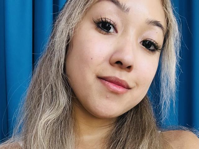 RenyLime's face, blonde hair, black eyelashes, pink lips, slight smile, light skin, blue curtain background, cute, no makeup, small beauty mark under right eye.