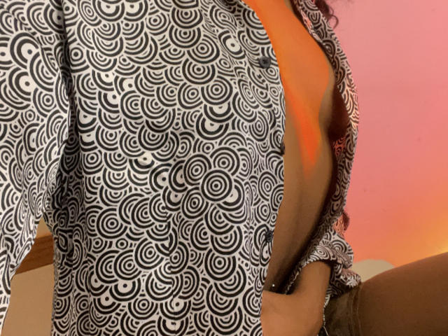 CelesteCharme's patterned shirt with black and white circular designs is open, revealing her tan skin and a hint of her brown pants. Her curly hair is partially visible.
