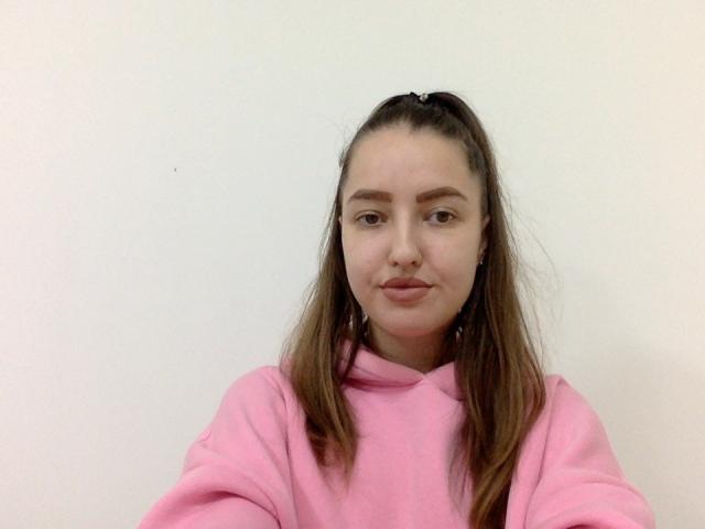 JessyHottie69 with pouty lips, brown eyes, and brown ponytail, wearing a pink hoodie, has small tits and a cute face, standing against a plain white wall.