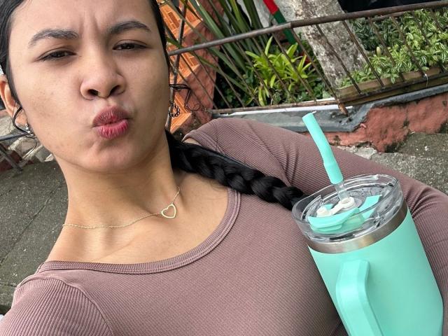 ChloeLauras with pouty lips, brown skin, black braid, pink top, heart necklace, holding turquoise drink with straw, outside.