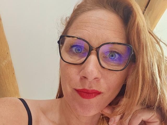 ApoloniaRed with red lipstick, blue eyes, and purple eyeshadow, wearing black glasses, has her hand on her chin, showing off her freckled face and reddish-brown hair.