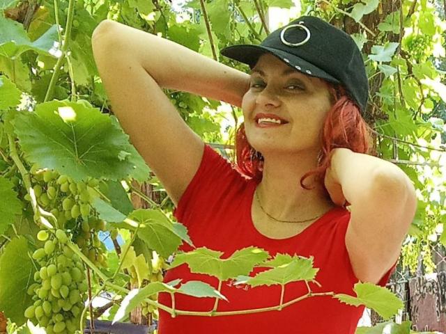 YanaFiesta, con cabello rojo, gorra negra y camisa roja, sonriendo, con los brazos levantados, mostrando los pezones, de pie en un viñedo de uvas, con hojas verdes y racimos de uvas en el fondo..